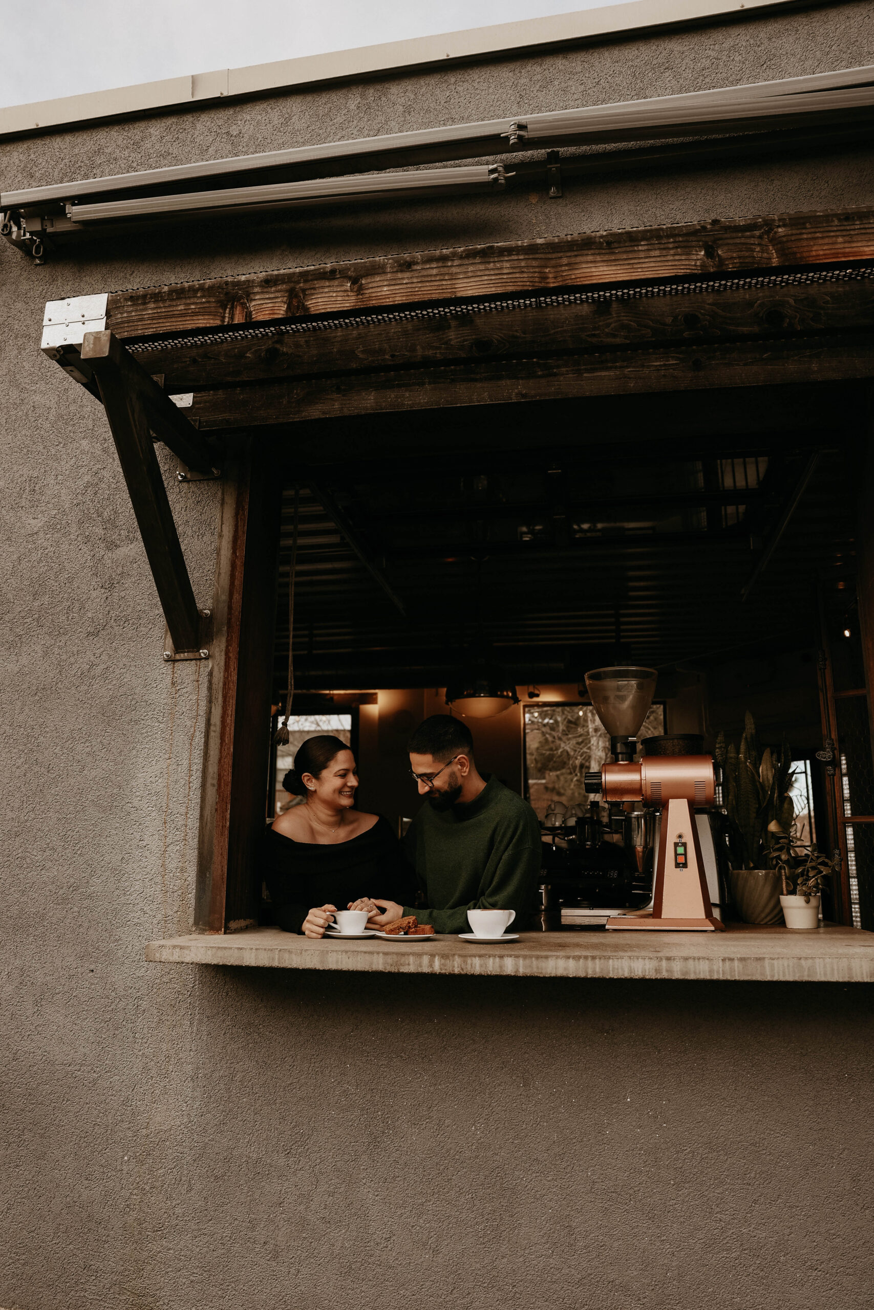 engaged couple at their engagement photo location, a coffee shop, standing in the window smiling with each other 