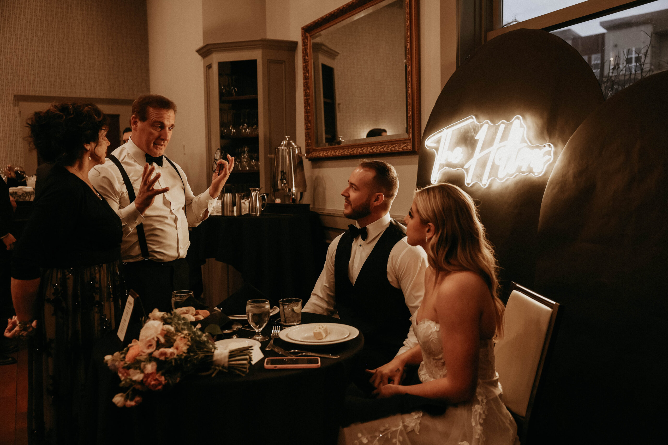 bride's dad talking to bridal couple while they sit at their reception table 