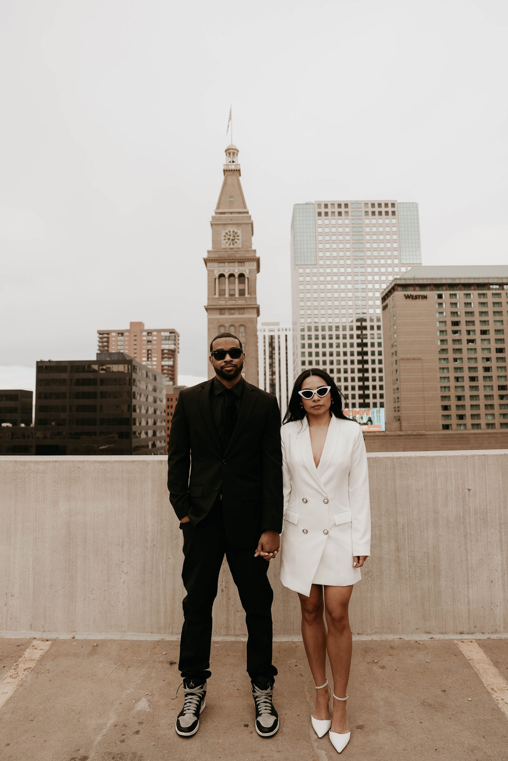 a newly engaged couple during a unique engagement shoot idea on a rooftop in Denver holding hands in formal attire wearing sunglasses 