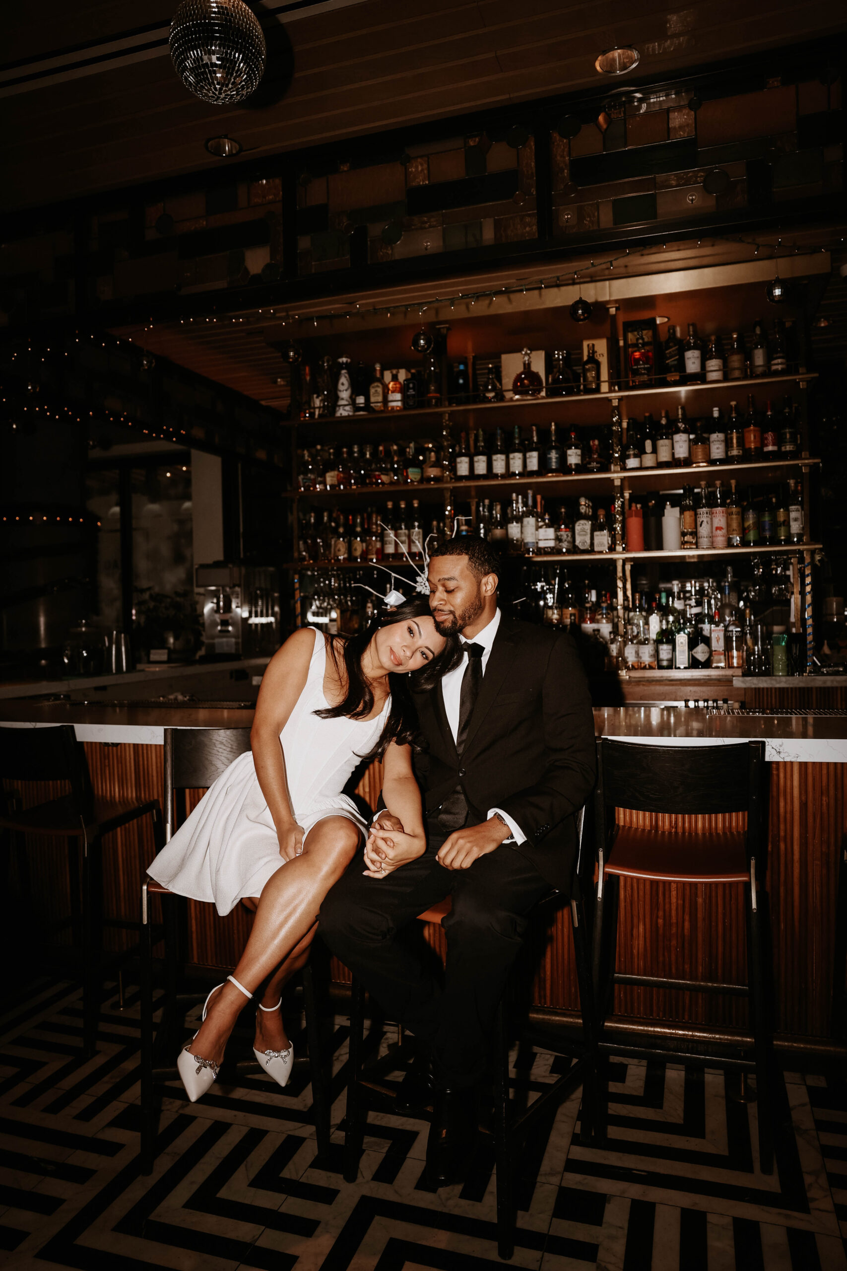 newly engaged couple sitting at a bar holding hands while one fiances puts her head on other fiances shoulder 