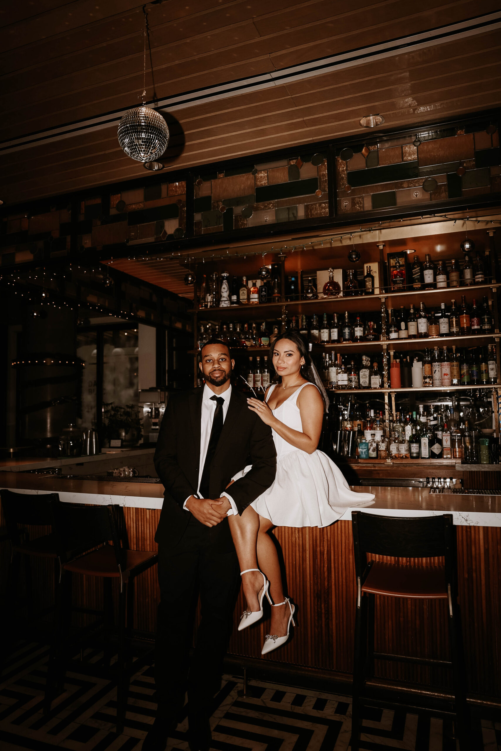 bride to be sitting on the bar and holding onto fiance standing next to her during a unique engagement shoot 