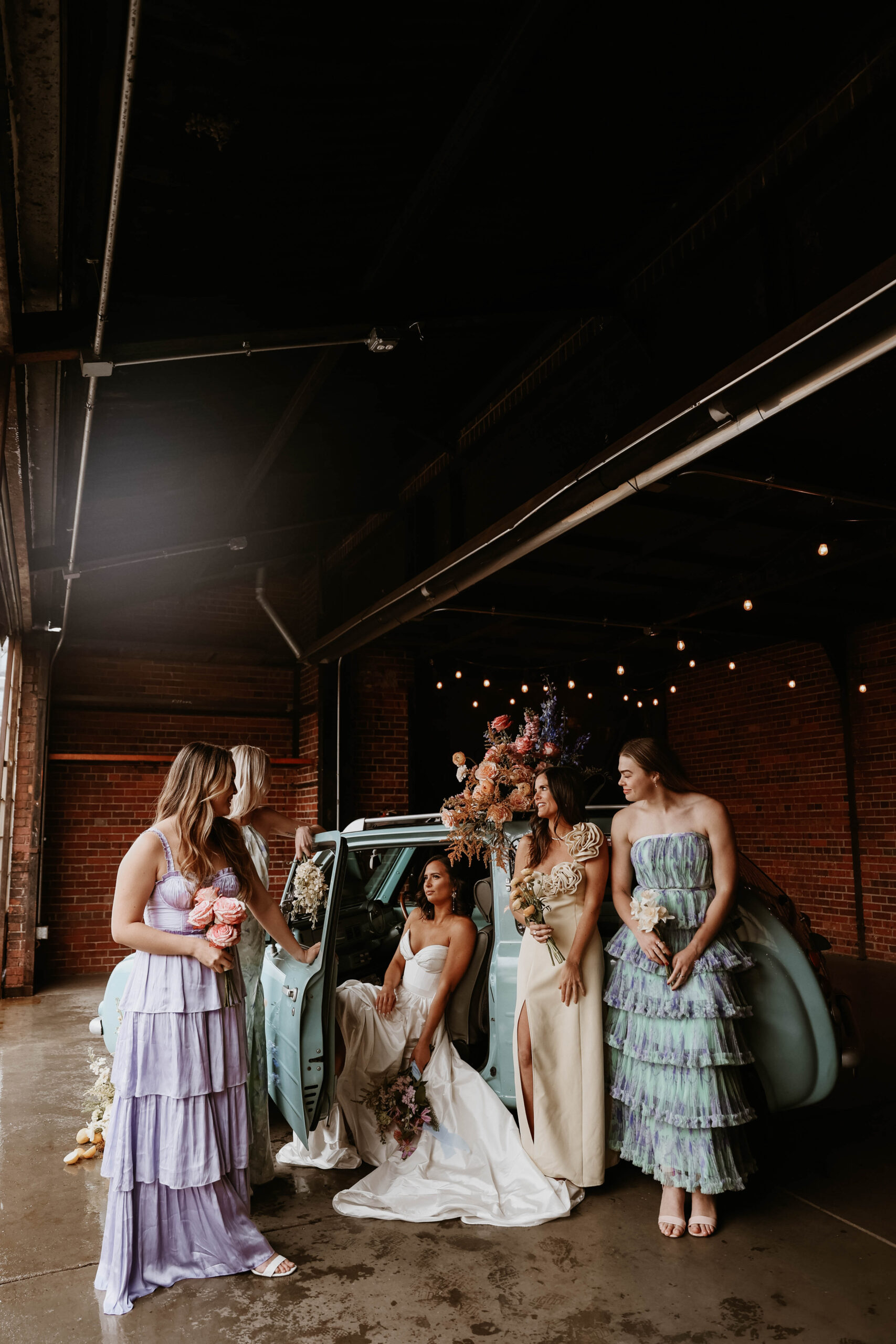 a bride sitting in s car while her bridesmaids stand around her during a colorful spring wedding 