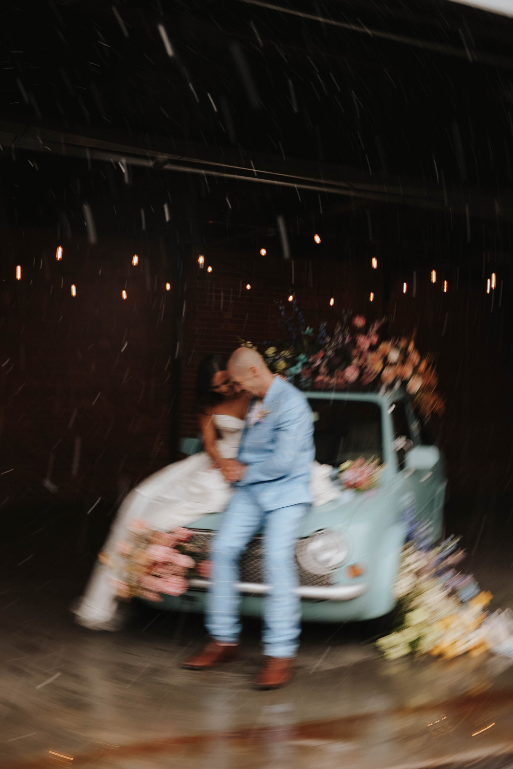 blurry photo of bride and groom sitting on the hood of a car during a colorful spring wedding 