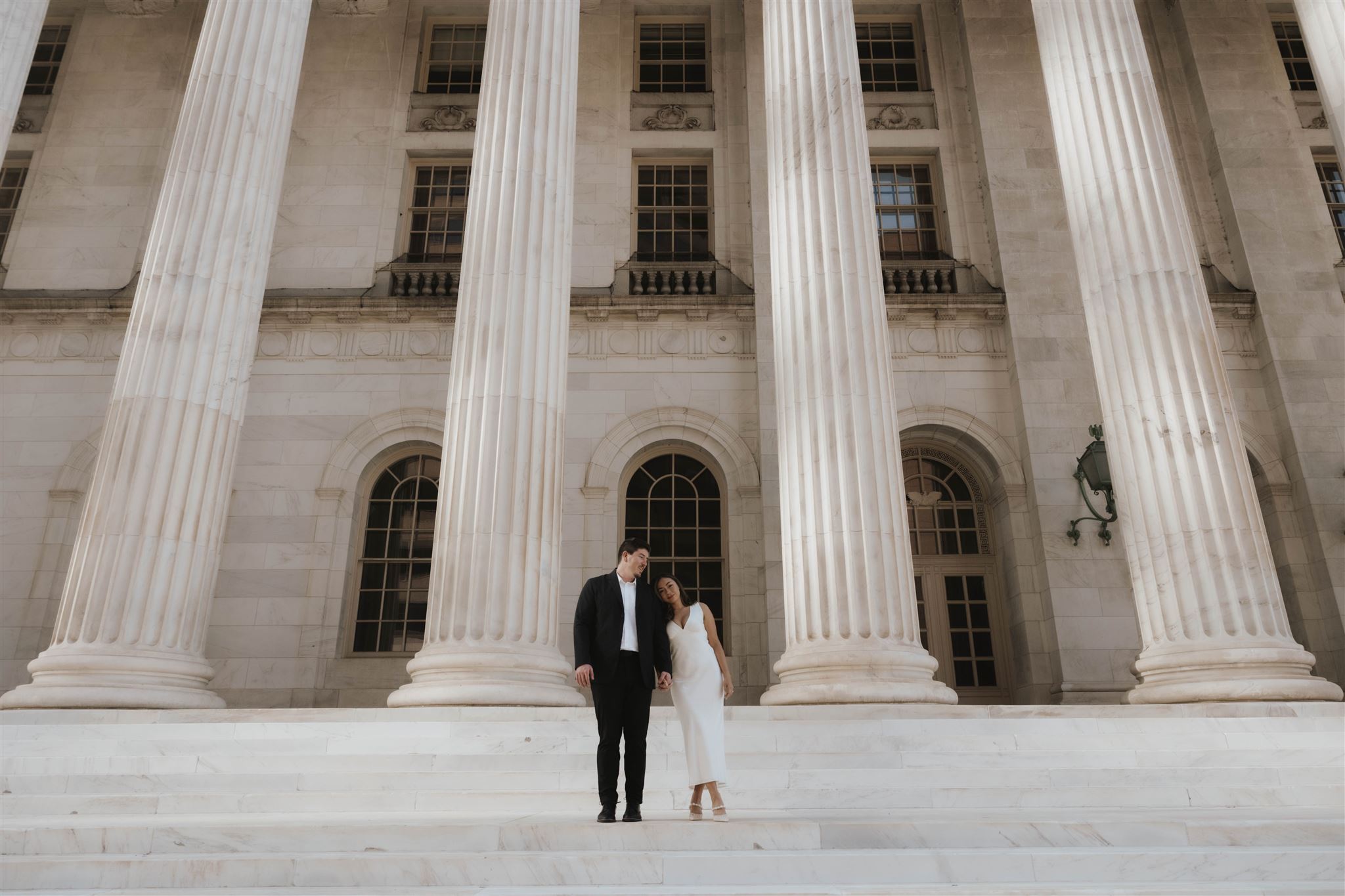 a woman resting her head on a man's shoulders as they stand in front of the courthouse for Elegant Engagement Photos