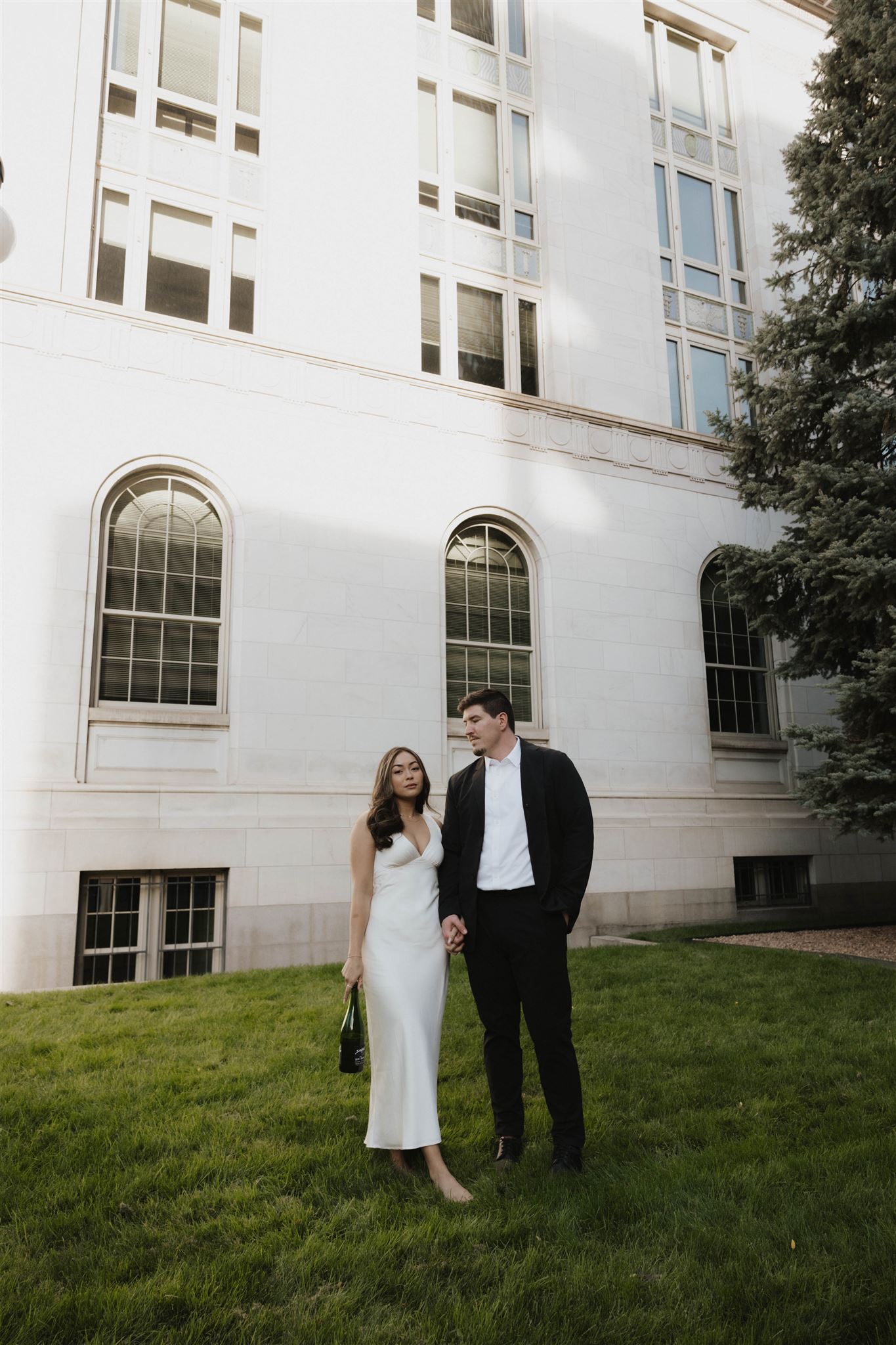 a man and woman standing in some grass with a bottle of champagne during their Elegant Engagement Photos