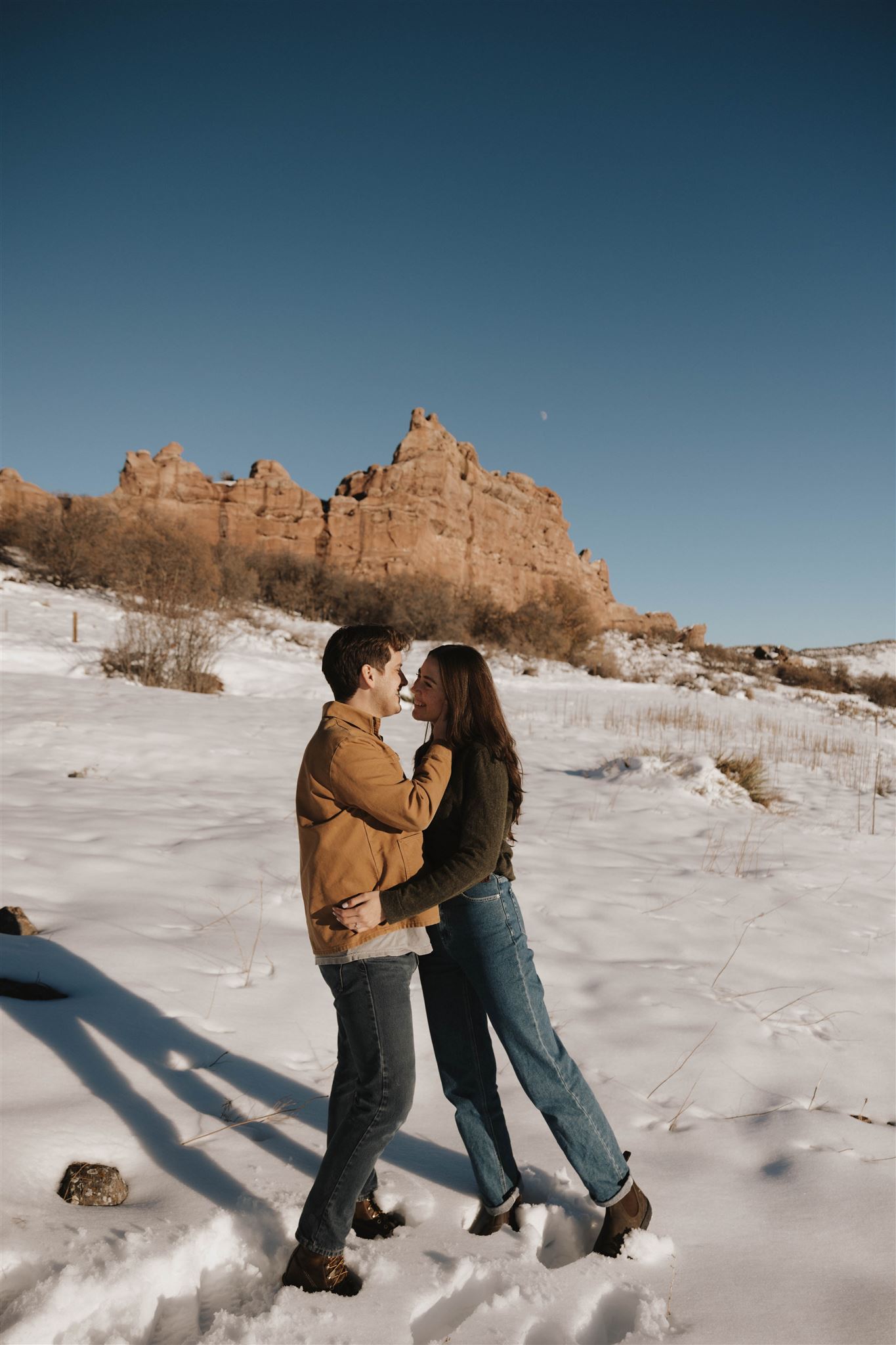 a man and woman smiling at each other while standing in the snow with red rocks behind them 