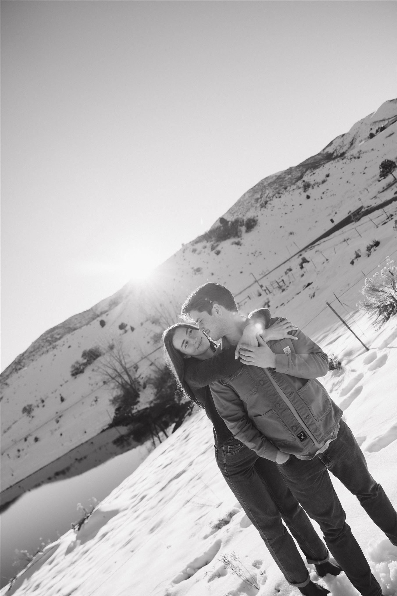 a woman holding onto a man from behind and smiling at him during their Snowy Engagement Photos