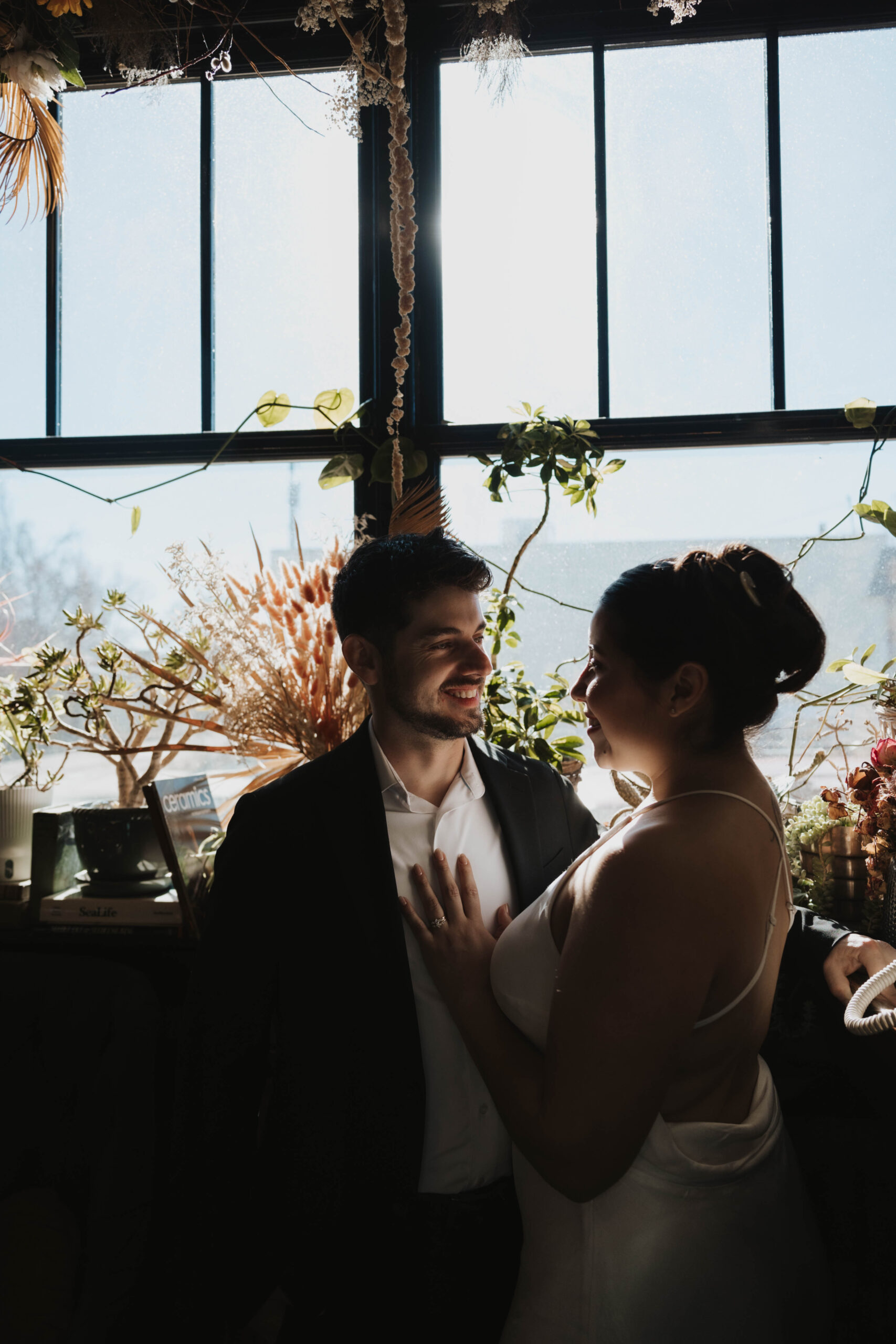 a man and a woman standing by a plant filled window for their engagement 